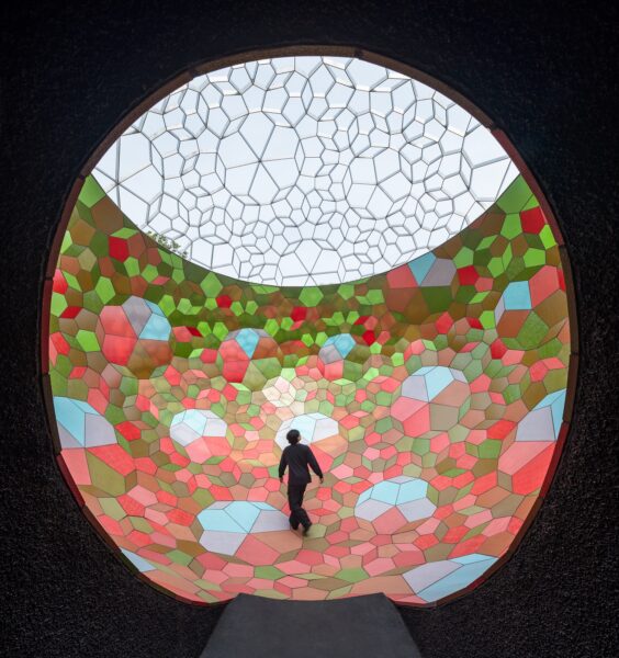 A dark tunnel leads to a big open space full of light and geometric colourful glass. A man is walking in the middle.