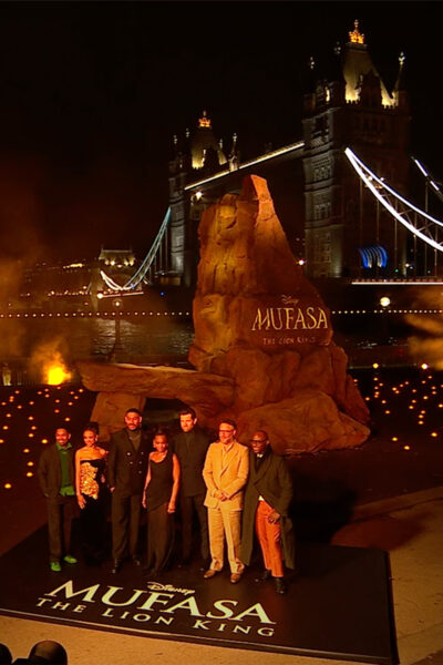 Celebrities stand in front a large scenic rock which has the word 'MUFASA' on it for a film premiere event.