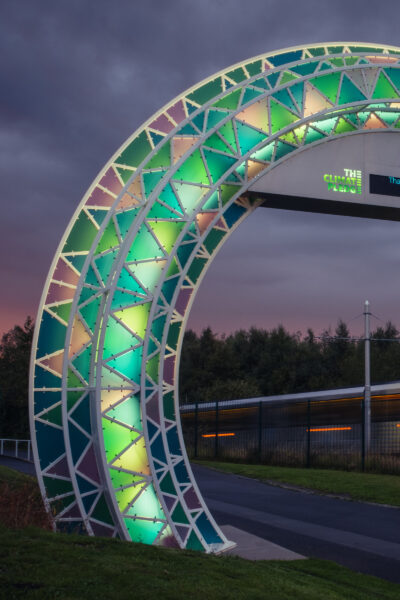 Illuminated arch lit up at night as a tram passes in the background.