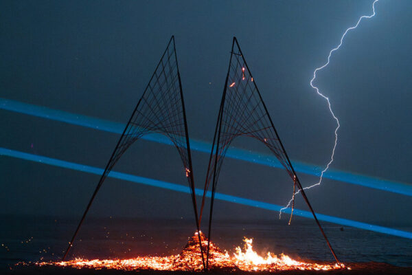 Night time shot of two metal structures on the beach. A fire burns below them and a lighting bold is striking the structure.