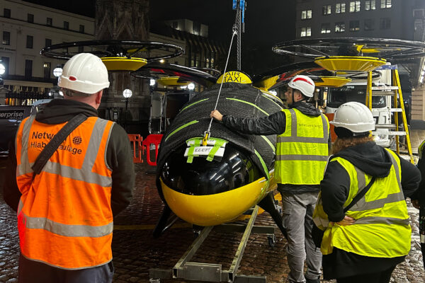 A flying taxi structure being installed by three people in high-vis vests and a crane.