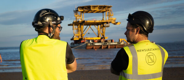 Two men in NEWSUBSTANCE high vis vests look out to see as an oil rig is shipped into shore.