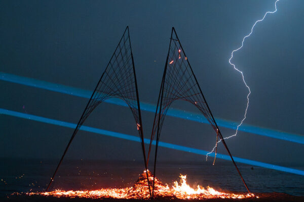 Night time shot of two metal structures on the beach. A fire burns below them and a lighting bold is striking the structure.