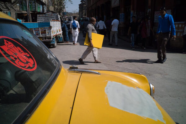 Yellow taxi with sqaure of missing yellow paint and in the background a man walks past the car holding a large yellow coloured sqaure