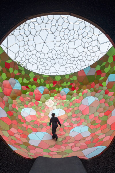 A dark tunnel leads to a big open space full of light and geometric colourful glass. A man is walking in the middle.